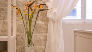 Une pièce avec un mur en pierre, une chaise blanche, un pouf en osier et un vase rempli de fleurs.