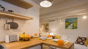 Une cuisine moderne avec un plan de travail en bois, un grille-pain et un panier de fruits. On y trouve un coin repas avec une table dressée pour deux personnes, comprenant des assiettes, des tasses et un verre de jus d’orange. Un canapé blanc avec un coussin coloré est visible à l’arrière-plan.