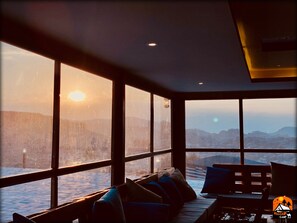 A room with large windows, a wooden bench, and a table with a lamp.