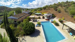 A pool area with a clear blue pool, surrounded by lounge chairs and umbrellas, set against a backdrop of hills and trees. - Fazendinha (Tavira)