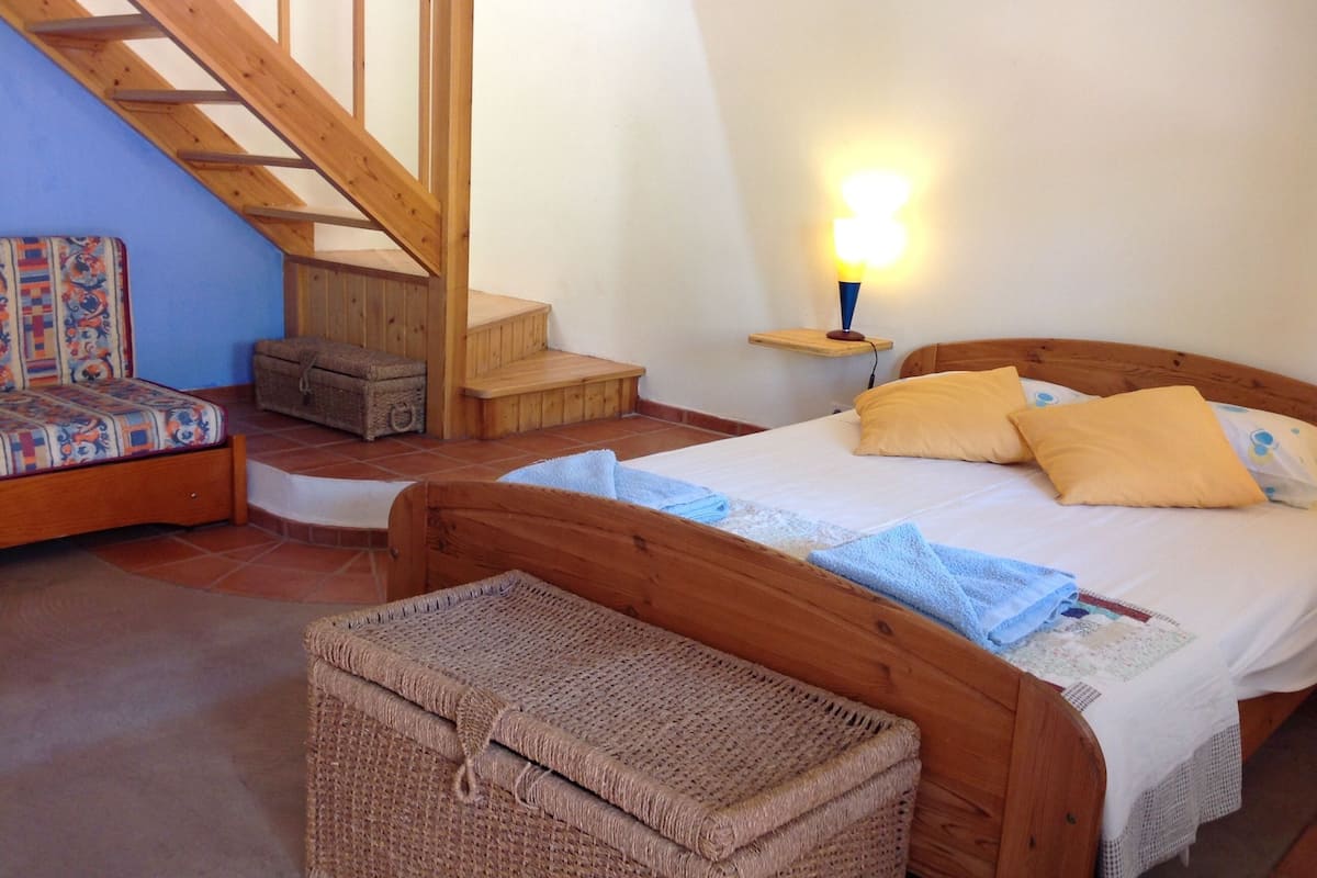 A bedroom with a wooden bed, a nightstand with a lamp, a staircase, and a small chest of drawers.