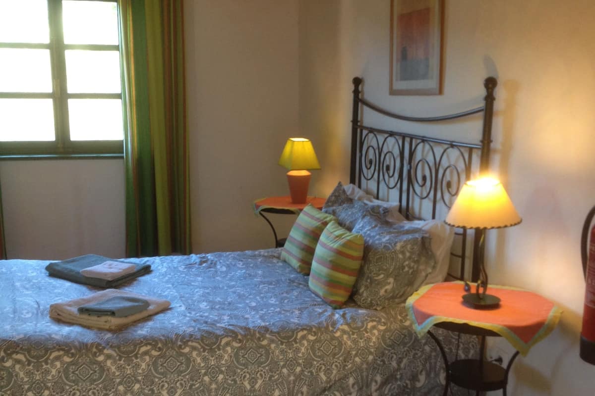 A bedroom with a bed, two bedside tables, a window with curtains, a lamp, and a painting on the wall.