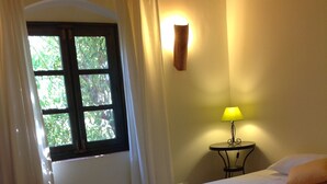 A hotel room with a bed, a window with curtains, a bedside lamp, and a view of greenery outside. - Fazendinha (Tavira)