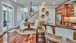 A spacious kitchen with wooden cabinets, a central island, and a dining area with white chairs and a woven rug. - Luxury Old Seagrove, BeachComer, Walk to Beach and Seaside, 3 King BRs, Private Heated Pool (Seagrove Beach)