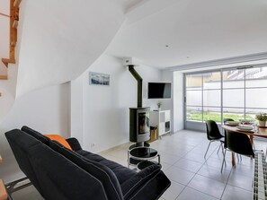 Un salon moderne avec un canapé noir, un téléviseur fixé au mur et une grande fenêtre donnant sur l’extérieur.