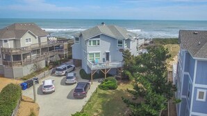 A beachfront property with a two-story house, a covered porch, and a parking area with several cars. - Serendipity - Oceanfront, Duck, 5 BR, 4.5BA, Private Pool & Hot Tub (Duck)