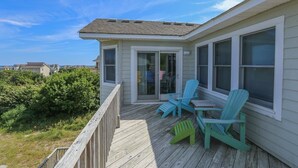 A covered deck with two chairs and a table, overlooking a grassy area with dense bushes. - Luzon - 5 BR, 4.5 BA, Oceanside home in Duck w/Private Pool (Duck)