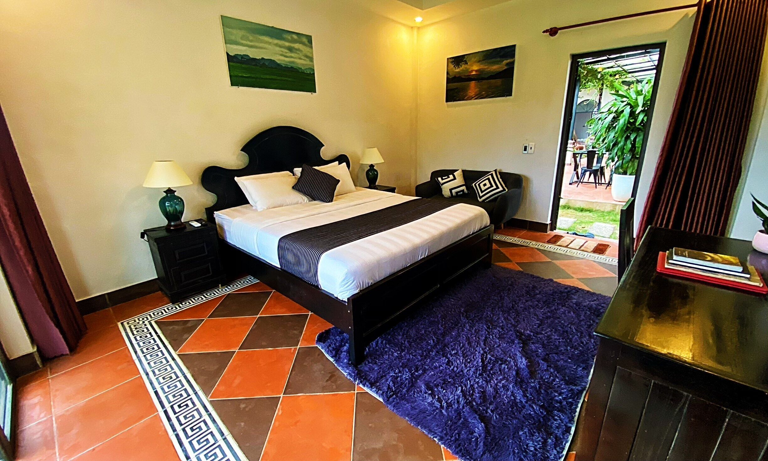 Habitación de hotel con cama, dos mesitas de noche con lámparas, una alfombra azul y vistas al exterior.