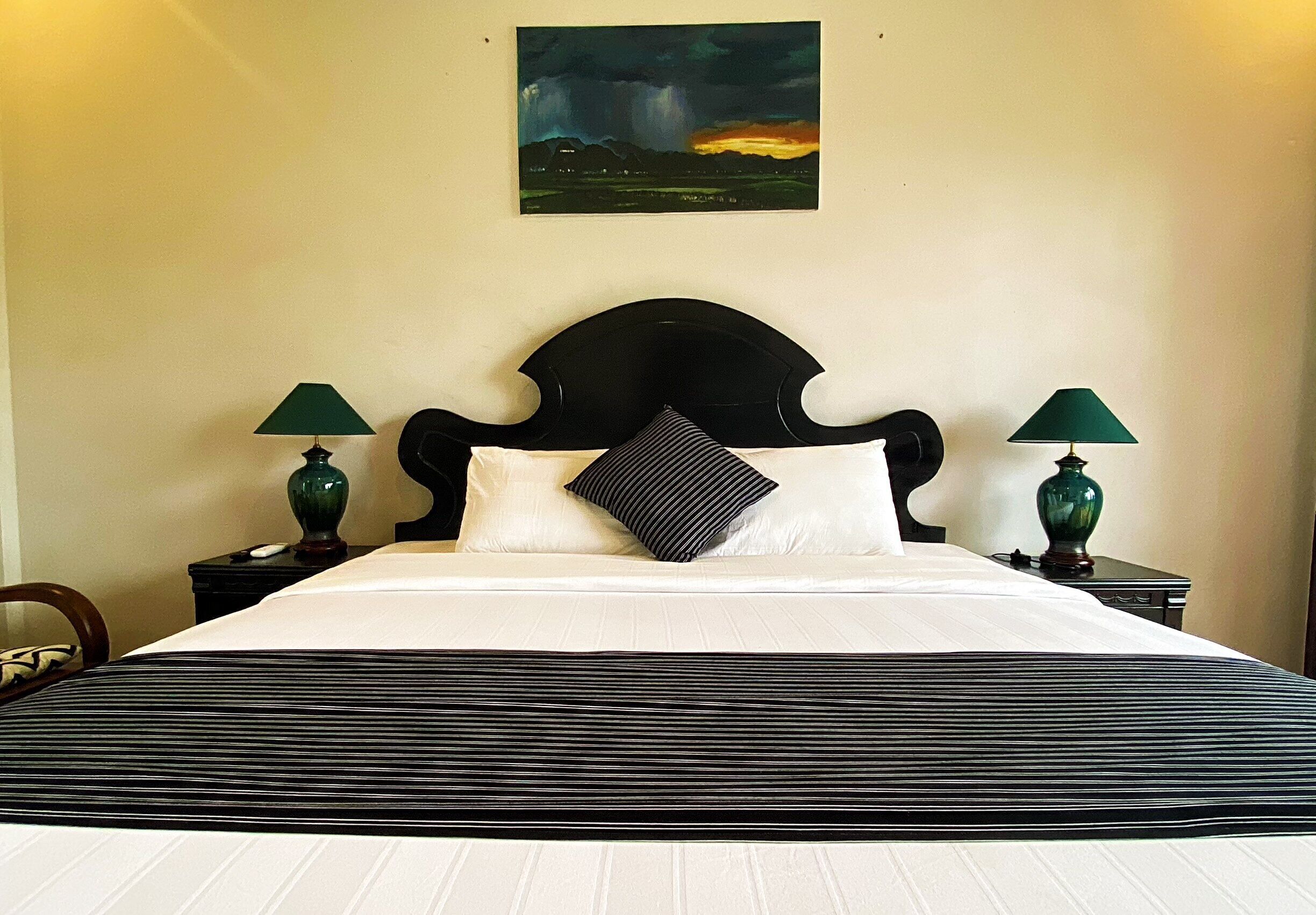 A neatly made bed with a striped bedspread, two green lamps on nightstands, a black headboard, and a painting of a stormy landscape on the wall.