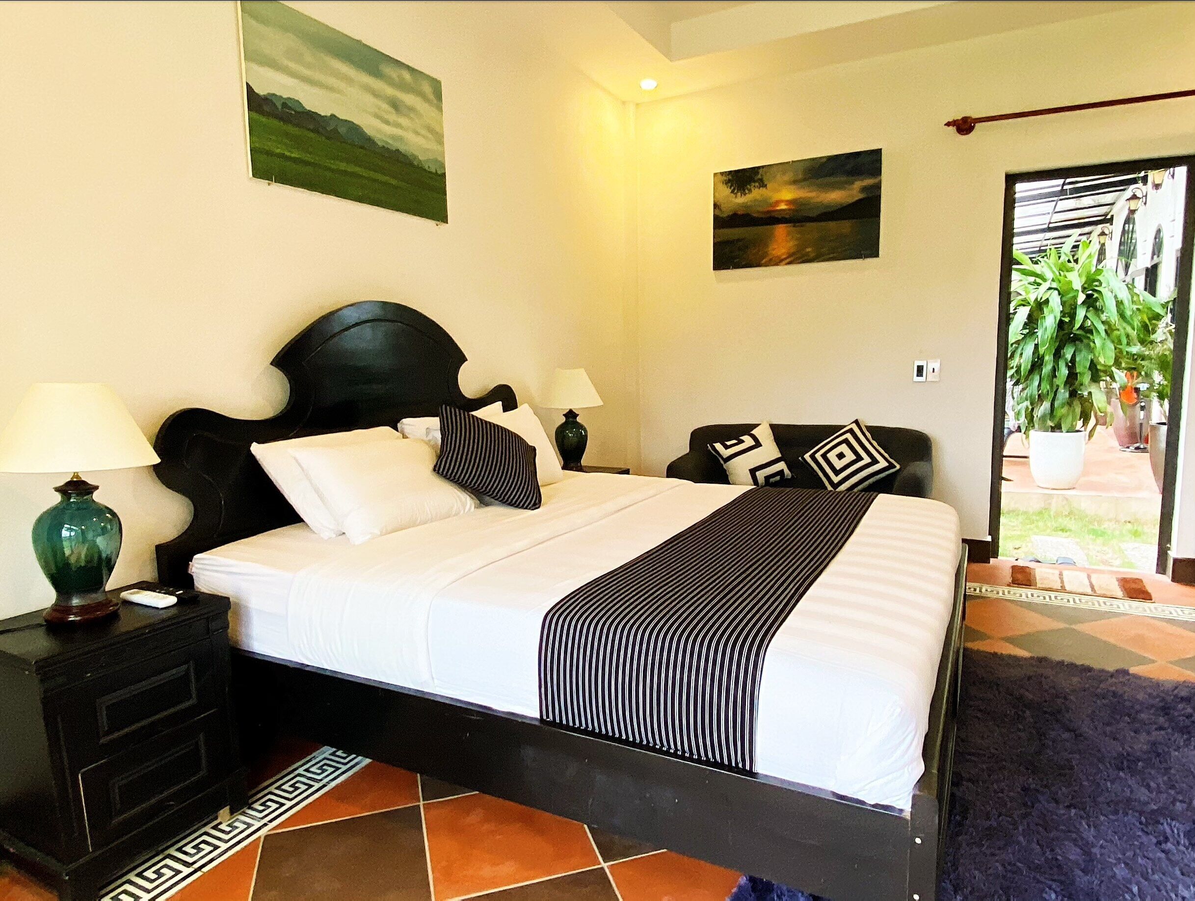 A neatly made bed with a black headboard, white bedding, and striped throw. There are two bedside tables with lamps, a small nightstand, and a decorative rug.