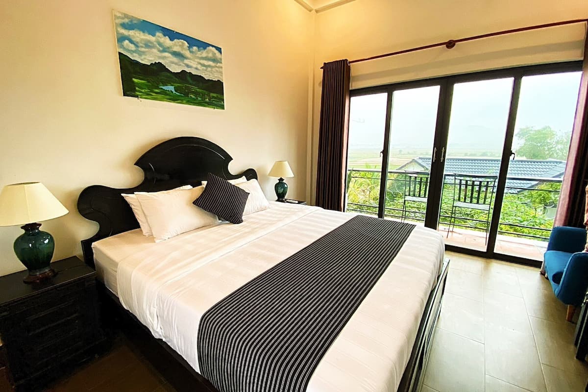 Habitación de hotel con una cama grande, dos mesitas de noche con lámparas, un cuadro en la pared y un balcón con vistas.