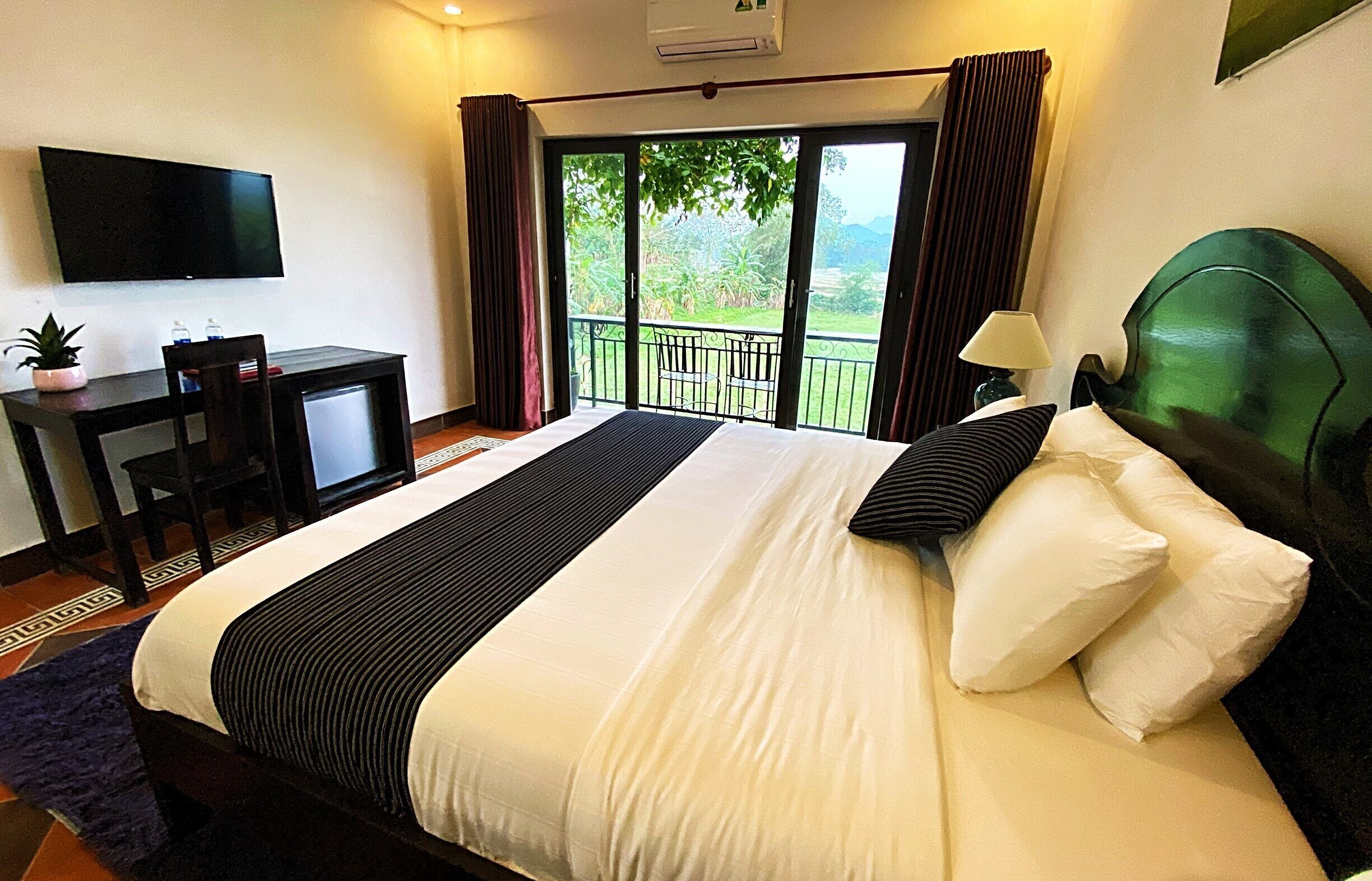 Habitación de hotel con una cama grande, un escritorio con televisor y un balcón con vistas a la vegetación.