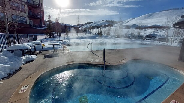 A hot tub with a view of a snowy mountain landscape, a fenced area with benches, and a building in the background. - Granby Ranch Ski Condo with King Bed & Bunkbed! (Granby)
