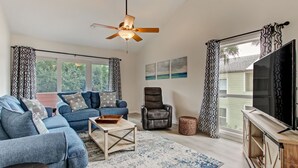 A modern living room with a blue sofa set, a leather armchair, a wooden coffee table, and a large flat-screen TV. - Renovated, Cozy 2Bd 2Ba, Steps from Private Beach & Pool! (St. Augustine)