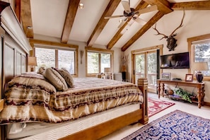 Une chambre avec un grand lit, des poutres en bois, un ventilateur de plafond, une tête de cerf fixée au mur et un téléviseur posé sur un meuble en bois.