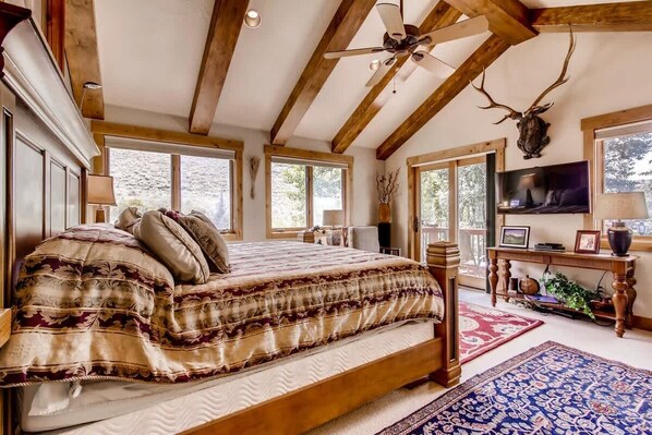 Une chambre avec un grand lit, des poutres en bois, un ventilateur de plafond, une tête de cerf fixée au mur et un téléviseur posé sur un meuble en bois.