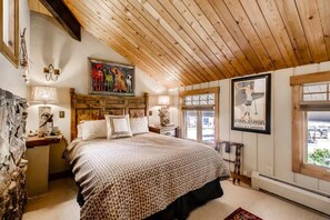 A bedroom with a wooden ceiling, a bed with patterned bedding, a painting above the bed, and a poster on the wall.