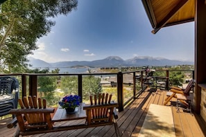 Una terraza de madera con dos sillas Adirondack, una mesa con un florero y vistas a montañas y un lago.