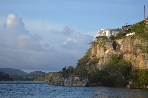 A coastal house with a balcony overlooking the sea, situated on a cliff with rocky terrain and sparse vegetation.