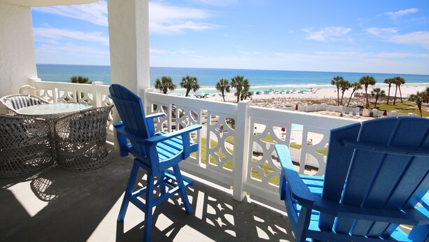 Un balcon avec des chaises bleues et une table donnant sur une plage bordée de palmiers et parsemée de parasols.