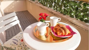 Une table ronde recouverte d’une nappe blanche, deux tasses blanches, un plateau avec des croissants et une chaise à motifs floraux.