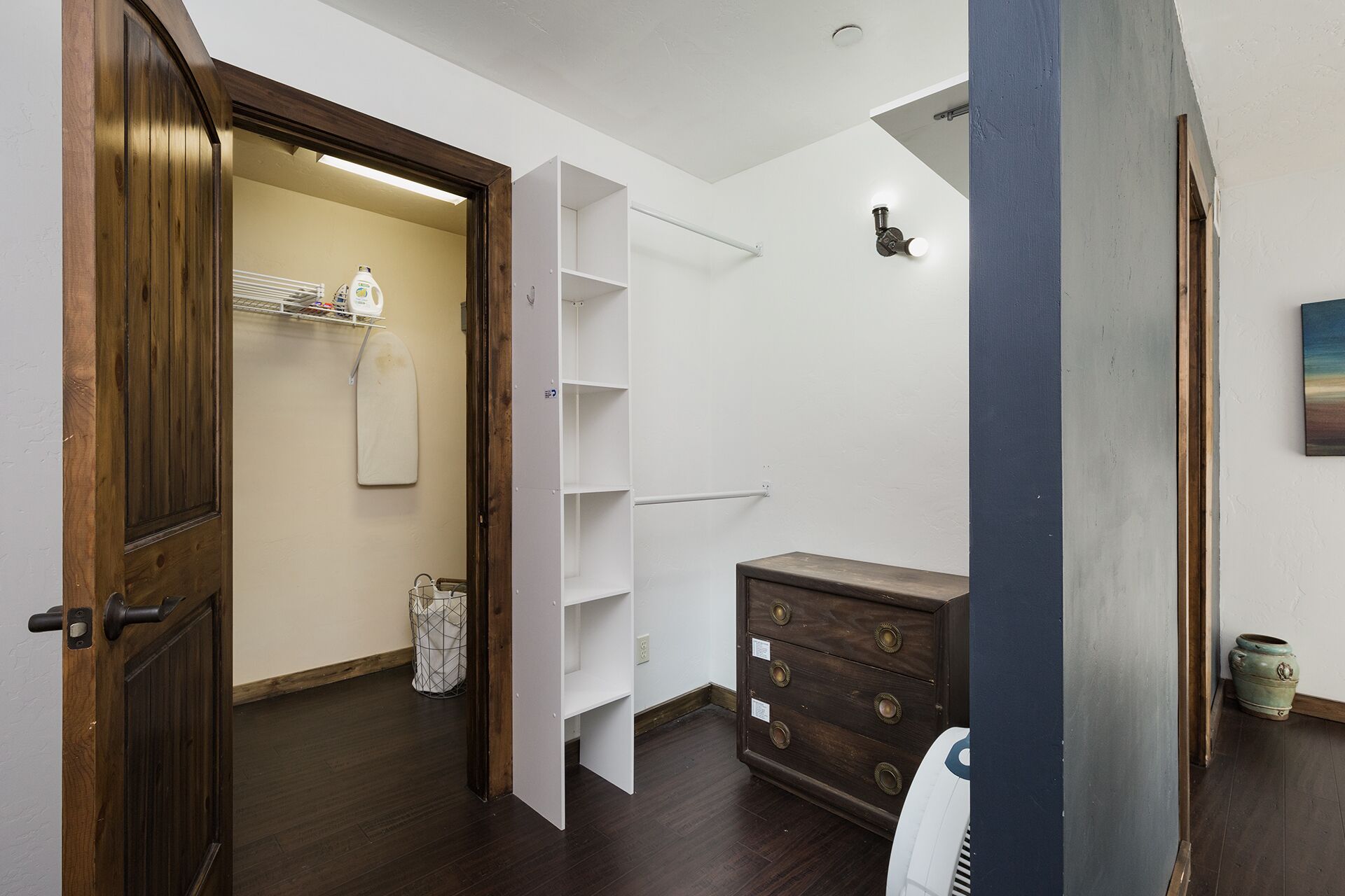 A room with a wooden door, a white shelf unit, a small wooden dresser, and a white fan.