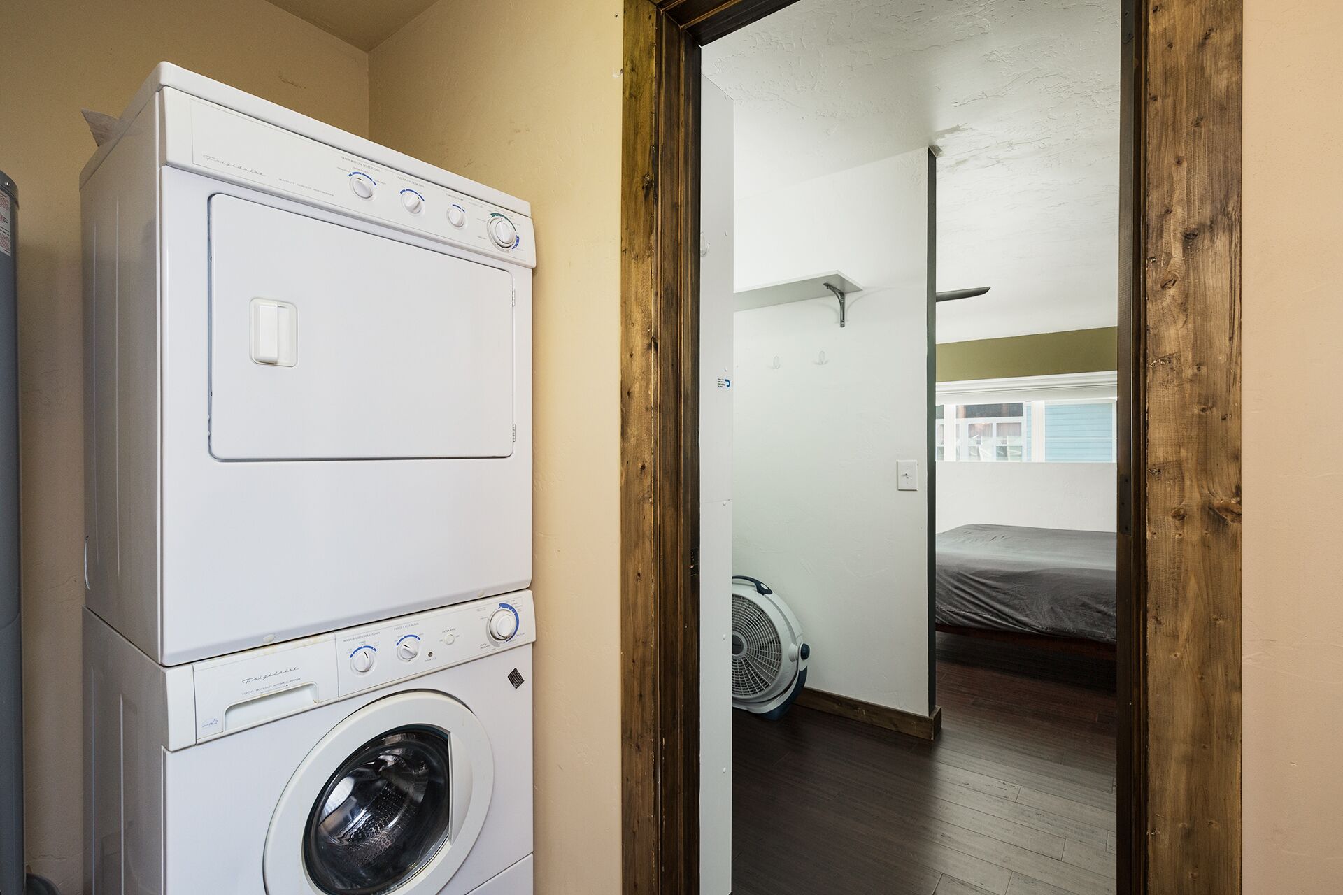 A laundry room with stacked washers and dryers, a wooden door, and a view into a bedroom with a bed and a fan.