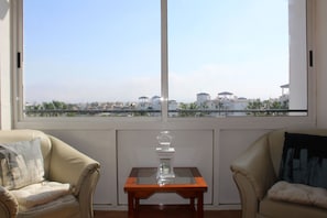 A room with a large window, two beige armchairs, a small wooden table with a glass vase, and a radiator below the window.