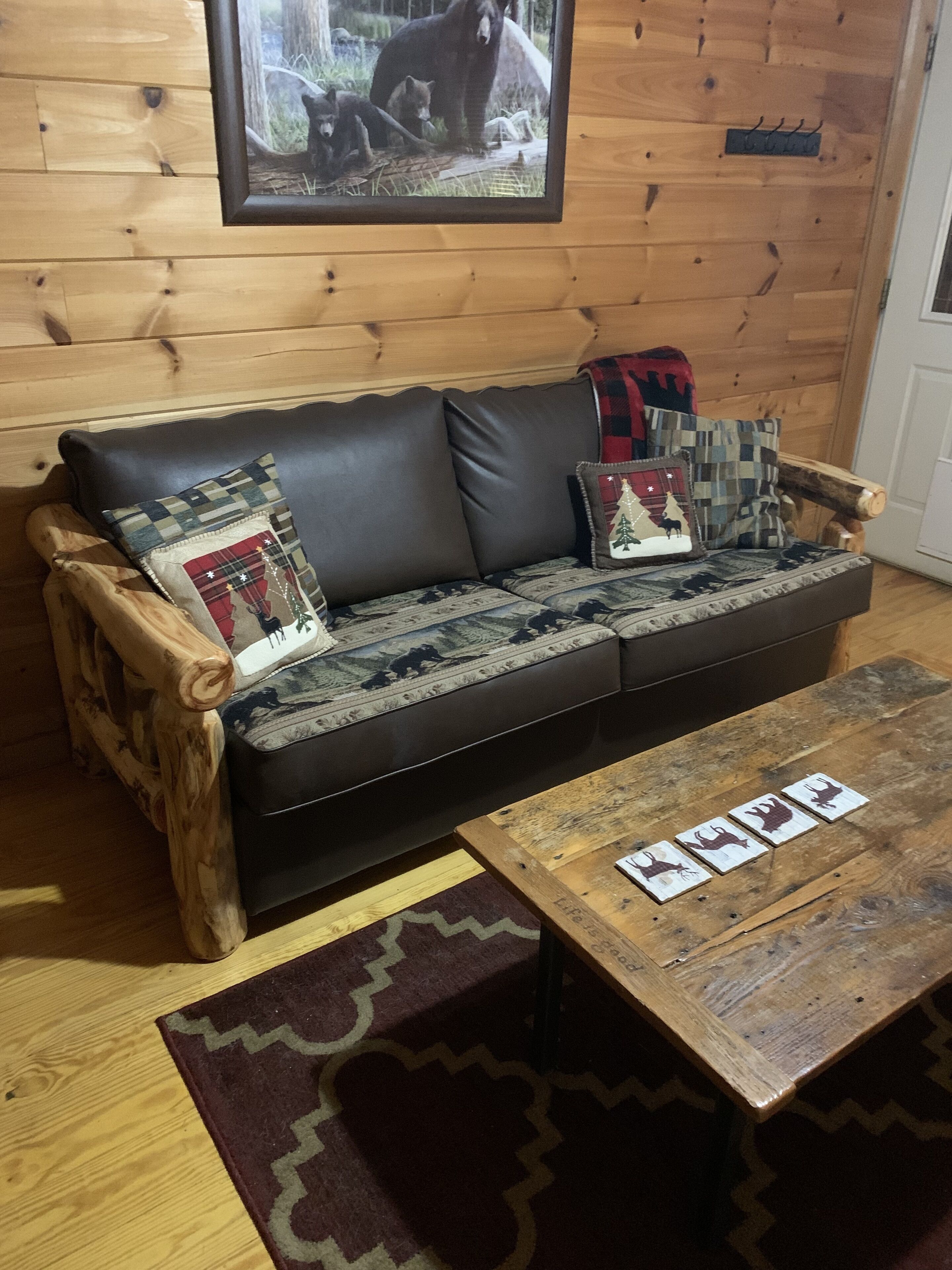Interior de una cabaña de madera con un sofá de cuero negro, dos almohadones decorativos, una mesa de centro de madera y una fotografía enmarcada de fauna silvestre en la pared.