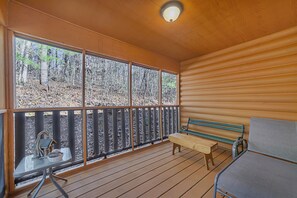 A wooden deck with a bench and a table, large windows with a view of a forest, and a ceiling light fixture. - Cozy mountainside cabin, seasonal views of Mt. LeConte & minutes from Dollywood. (Pigeon Forge)