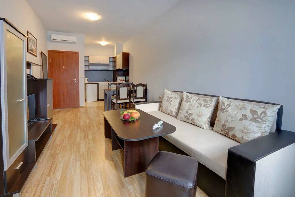A compact living space with a sofa, a dining table with fruit, a kitchenette, and a TV.