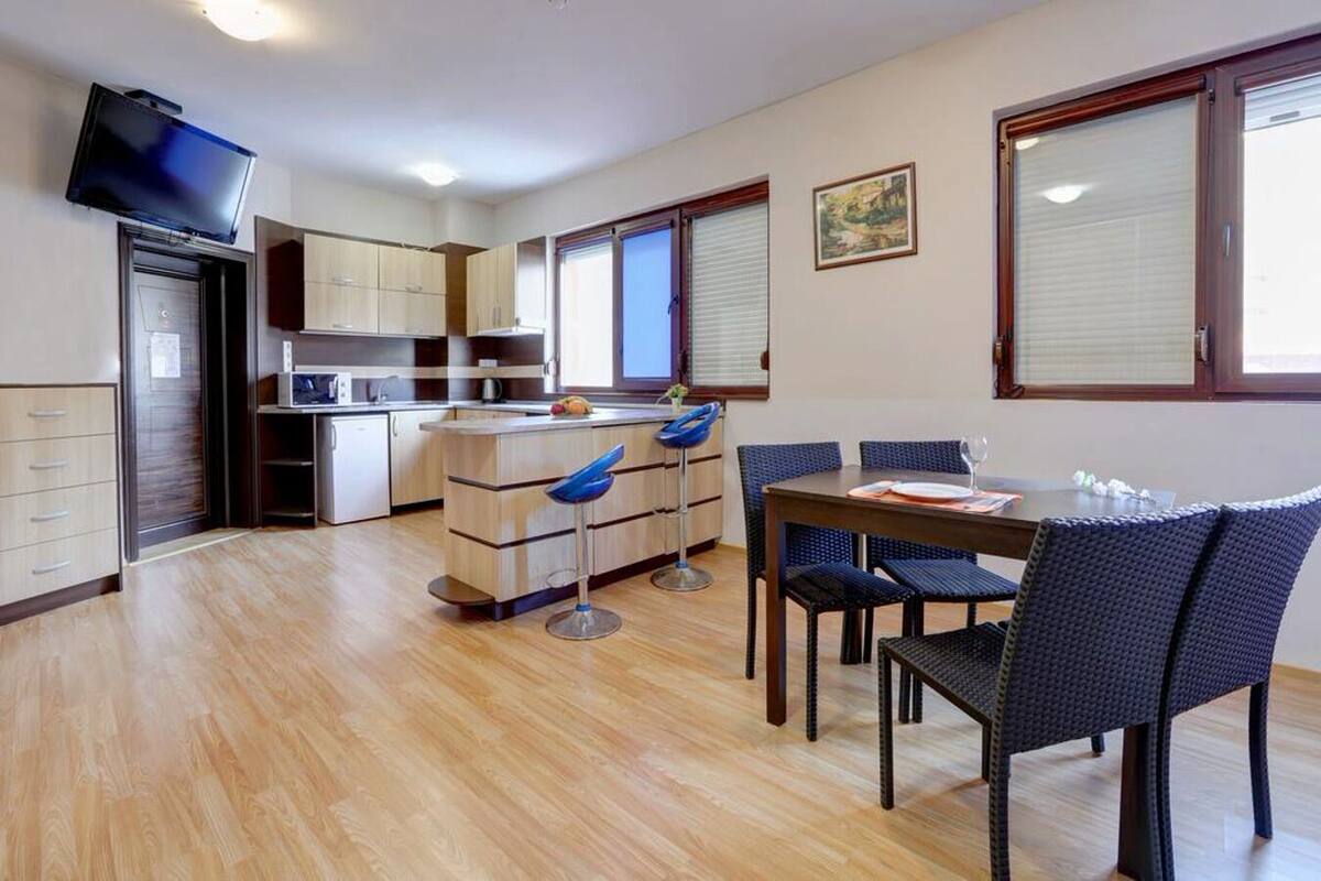 A compact kitchen with a dining area, a TV mounted on the wall, and a window with blinds.
