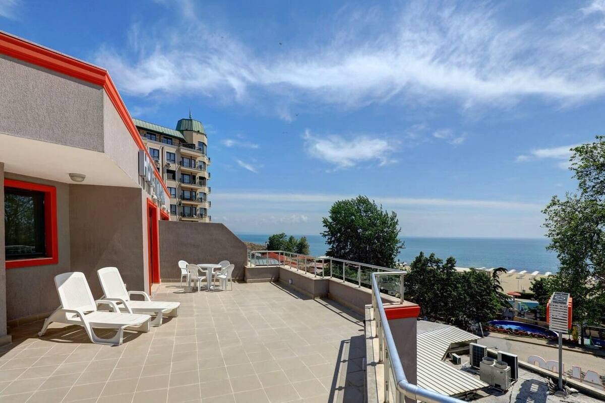 A rooftop terrace with white chairs and a table, overlooking a beach and a building with a green roof.