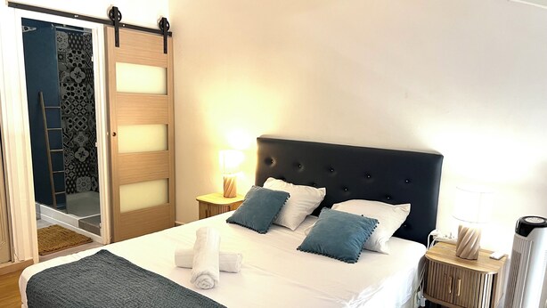 A neatly made bed with a dark headboard, two pillows, a blanket, and a towel on the side. There is a bedside table with a lamp and a small cabinet. - Cozy studio in Le Gosier 2 minutes from the beach (Le Gosier)