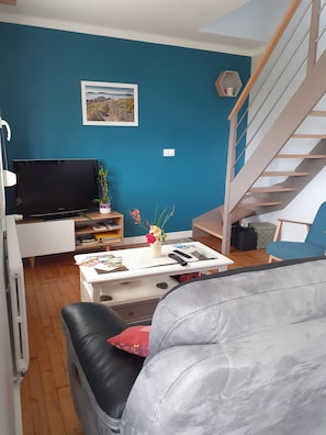 Un salon comprenant un canapé, une table basse ornée d’un vase de fleurs, un téléviseur à écran plat et un escalier.