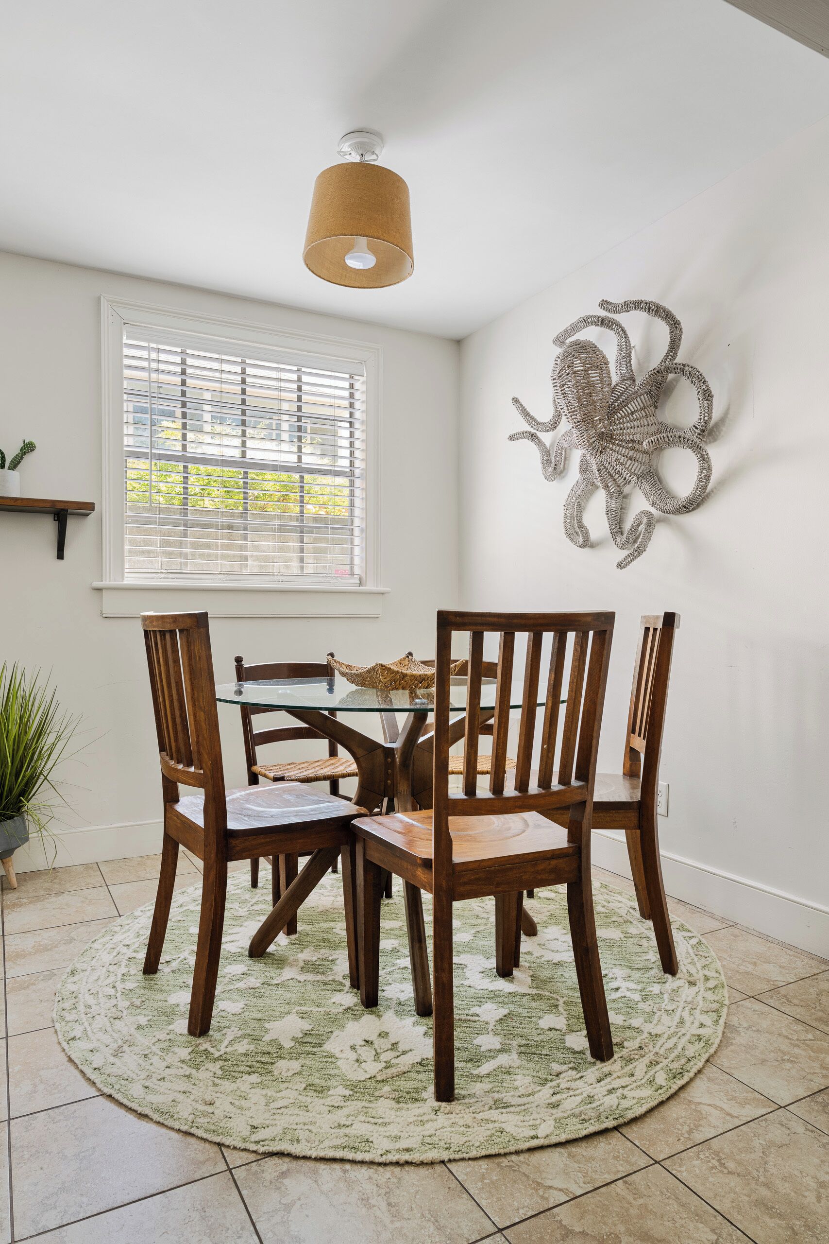 A dining area with a glass table, wooden chairs, a woven rug, a ceiling light fixture, and a wall-mounted decorative piece.