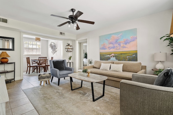 A living room with a sofa, armchairs, a coffee table, a ceiling fan, and a painting on the wall.