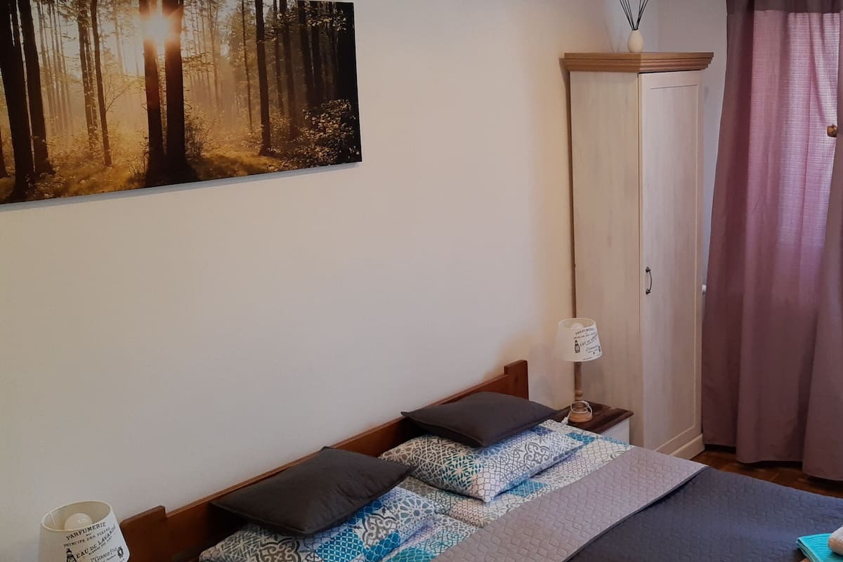 A bedroom with a bed, a nightstand, a radiator, and a window with curtains.