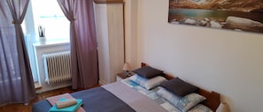 Une chambre à coucher avec un lit, une table de chevet, une lampe, un radiateur et une fenêtre avec des rideaux.