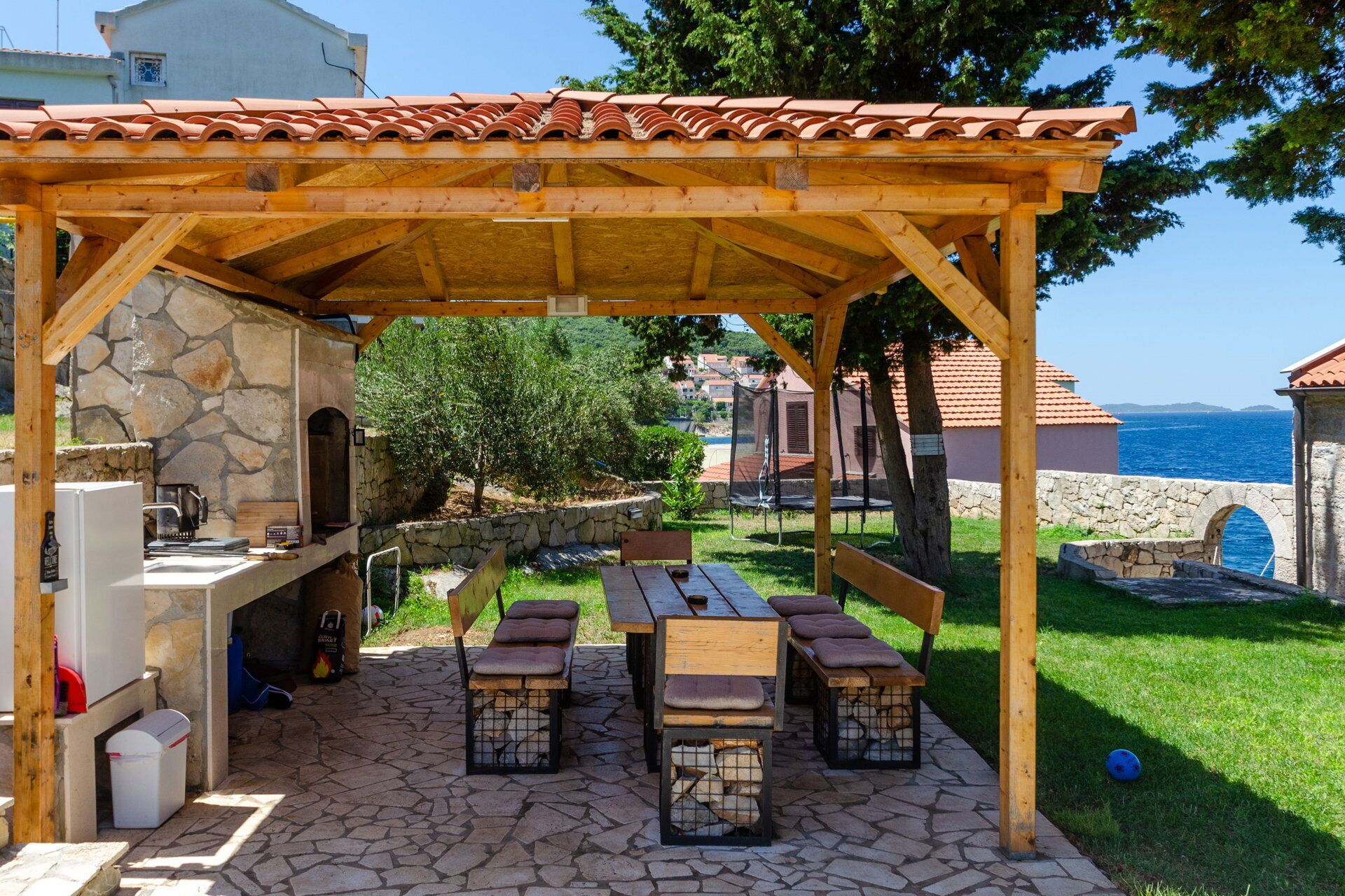 An outdoor patio with stone flooring, wooden benches, a grill, and a stone wall.
