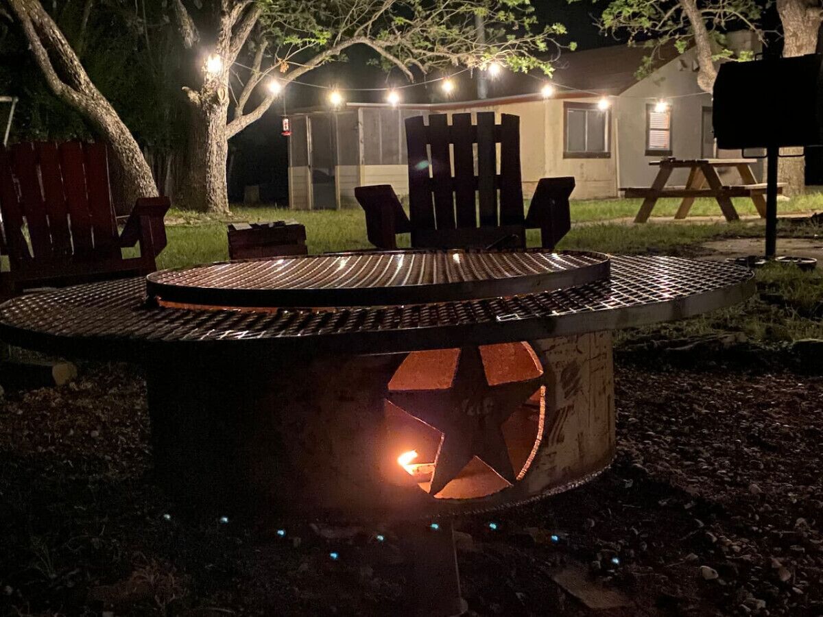 A circular outdoor fire pit with a metal grill, surrounded by chairs and a table.