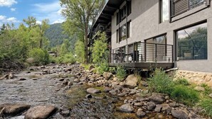 A modern building with a stone foundation, large windows, and a balcony overlooking a rocky stream.