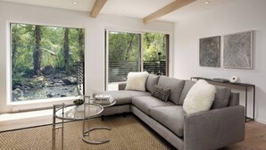 A modern living room with a grey sectional sofa, a glass coffee table, and a view of a forest through large windows.