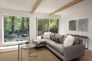 Un salon moderne avec un canapé d’angle gris, une table basse en verre et une vue sur une forêt grâce à de grandes fenêtres.