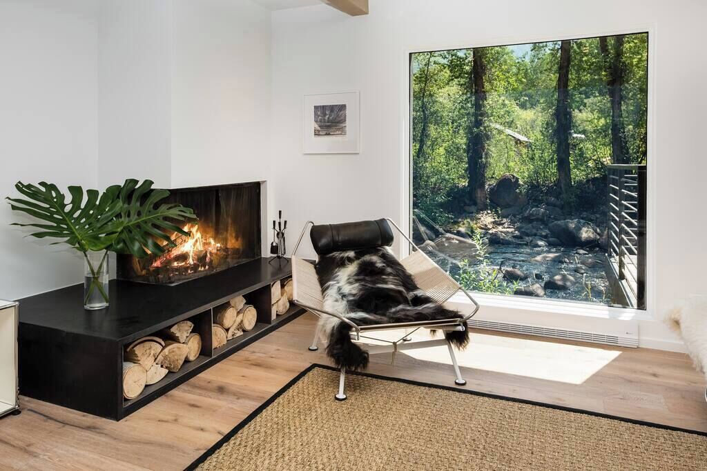Una sala de estar moderna con chimenea, un ventanal con vista a un bosque y un perro recostado en una silla.