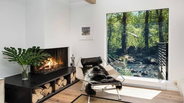 A modern living room with a fireplace, a large window with a forest view, and a dog lying on a chair.