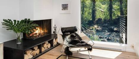 Una sala de estar moderna con chimenea, un ventanal con vista a un bosque y un perro recostado en una silla.