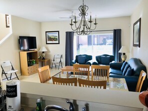 Uma sala de estar com televisão, lustre, mesa de jantar com cadeiras, sofá e janela com cortinas.