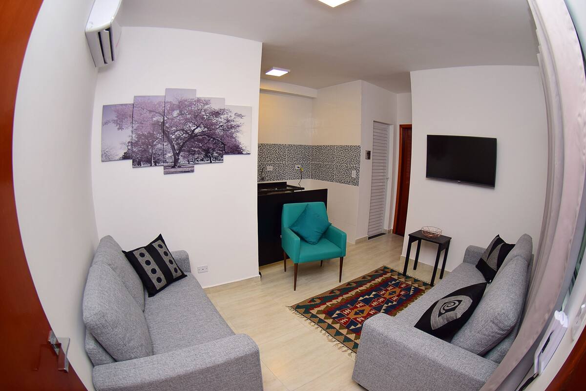 A living room with a grey sofa, teal armchair, and a flat-screen TV mounted on the wall.
