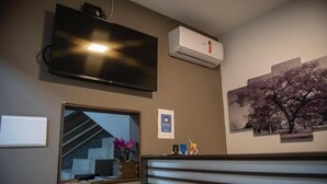 Une chambre équipée d’un téléviseur fixé au mur, d’un appareil de climatisation, d’une fenêtre donnant sur un escalier et des fleurs, ainsi que d’un tableau représentant un arbre.